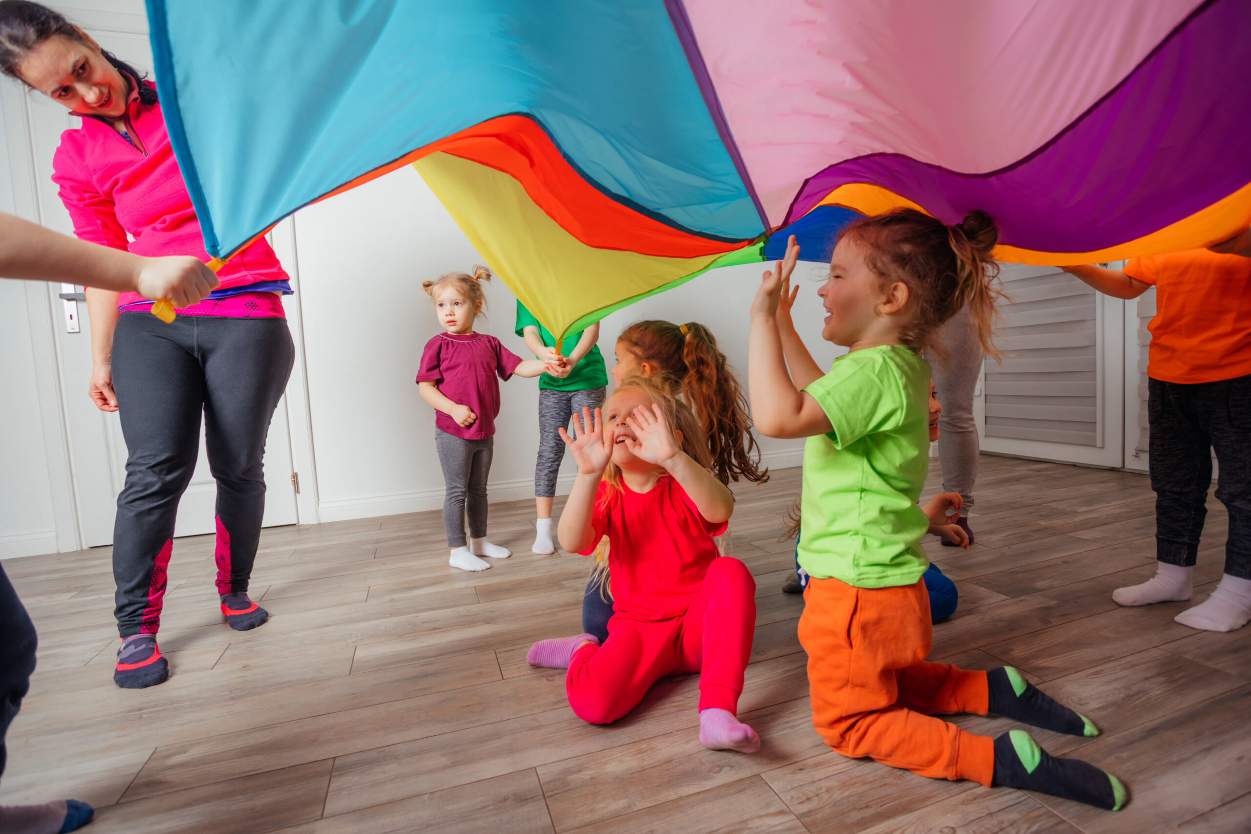Close up view of happy and excited kindergarten children under huge rainbow cover supported by other children and teachers. Children enjoying games with colorful canopy.